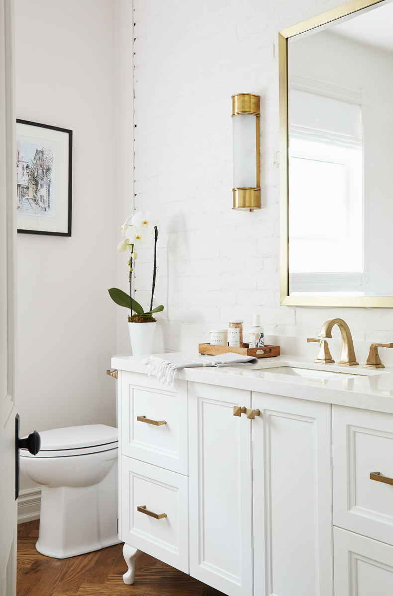 gorgeous bathroom with toilet in corner, white vanity with gold hardware, gold faucet and mirror with wall sconce above 
